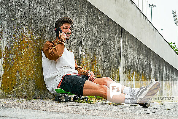 Junger lässiger Mann sitzt im Freien auf einem Skateboard und benutzt ein Telefon