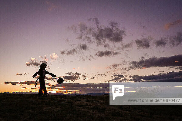 A young girl a twirls a halloween bucket on top of a hill at sunset