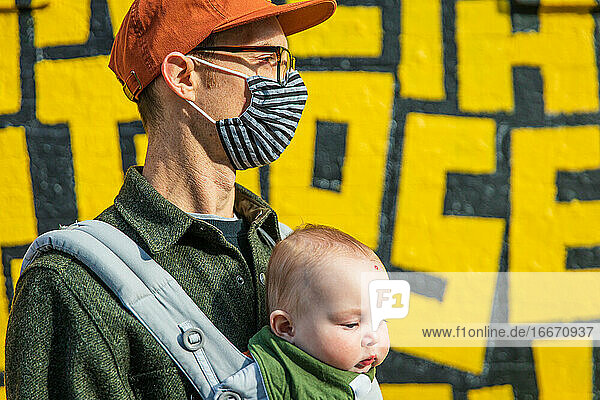 Vater mit Gesichtsmaske trägt süße Tochter  während er während des Ausbruchs des Coronavirus an einer Graffiti-Wand steht