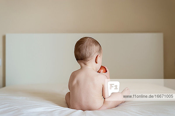 Nacktes Baby sitzt auf dem Bett auf dem Rücken und hält einen Apfel