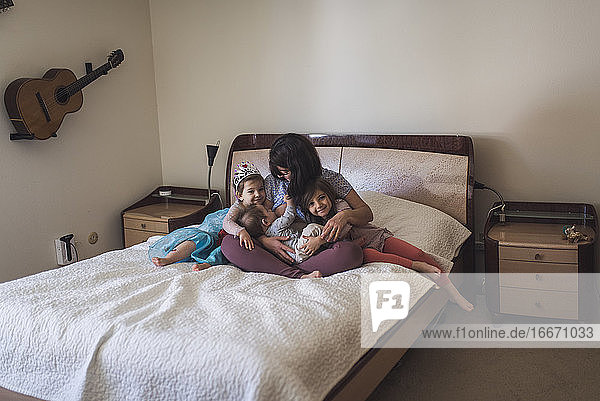 MId-30's mom nursing baby on bed and hugging 4- and 6-yr old daughters