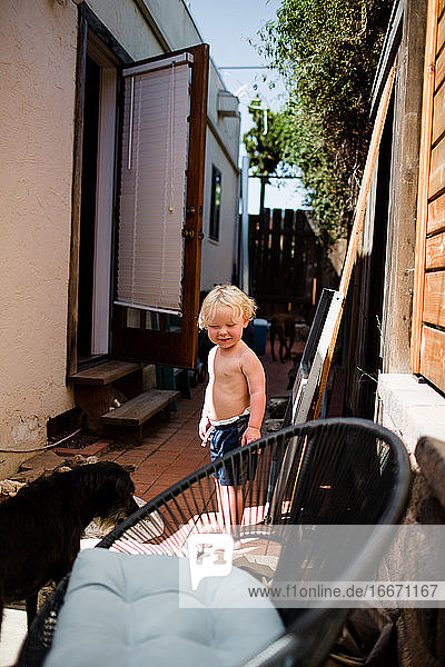Shirtless zwei Jahre alt stehend auf Seite des Hauses lächelnd auf Hund