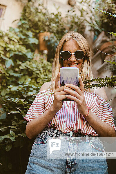 Glückliche Frau mit Sonnenbrille schaut auf ihr Smartphone in der Natur