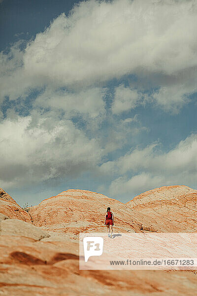 woman hiker walks up a red rock hill towards blue cloudy skies in utah