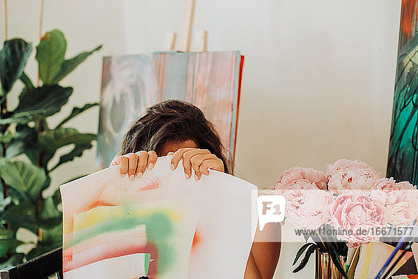 Eine Malerin versteckt sich in ihrem Atelier hinter einem Blatt Papier