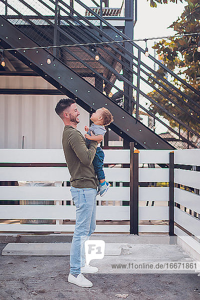Dad laughing while holding boy in front of white fence and black steps