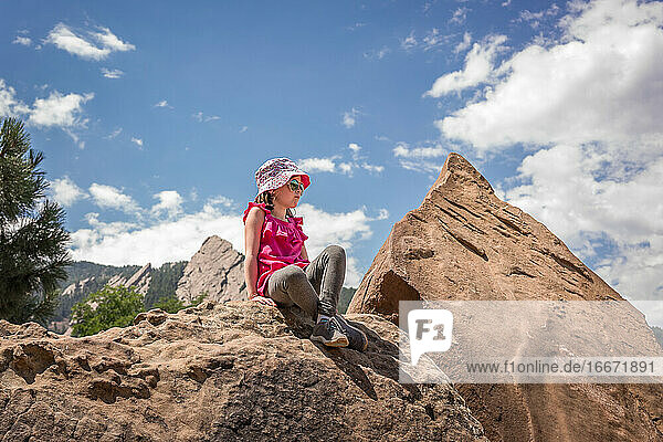 junges Mädchen mit Hut sitzt auf einem Felsen mit Bergen im Hintergrund nach einer Wanderung