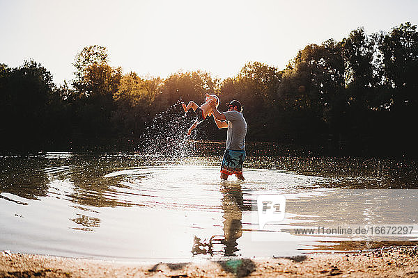 Vater hält kleines Kind in der Luft am See und spritzt Wasser
