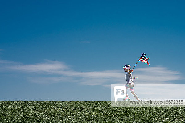 junges Mädchen läuft über einen Grashügel und schwenkt 2 amerikanische Flaggen