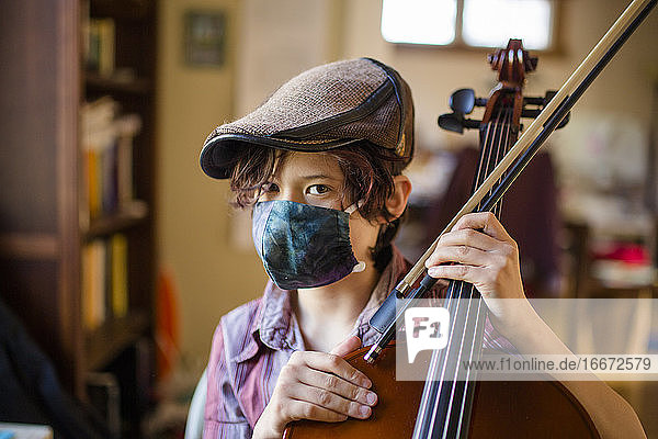 Ein ernster Junge mit direktem Blick  der eine Maske und eine Wollmütze trägt  hält ein Cello