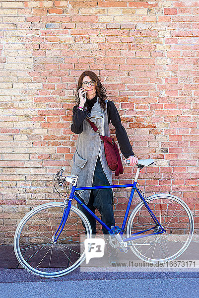 Lächelnde junge Frau mit Fahrrad  die sich an die Wand lehnt und ein Handy benutzt