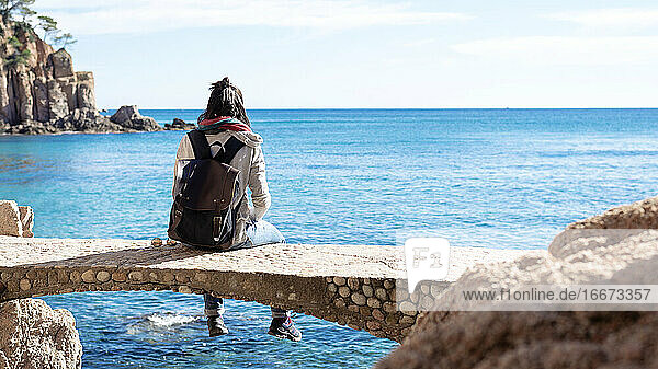 Rückansicht einer Frau  die sich vor dem blauen Meer entspannt  auf einer Brücke sitzend