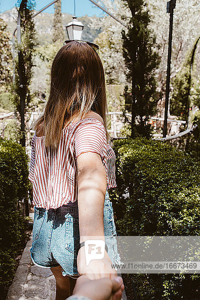 Rückenansicht einer blonden Frau  die Hände hält und in einem Naturpark spazieren geht