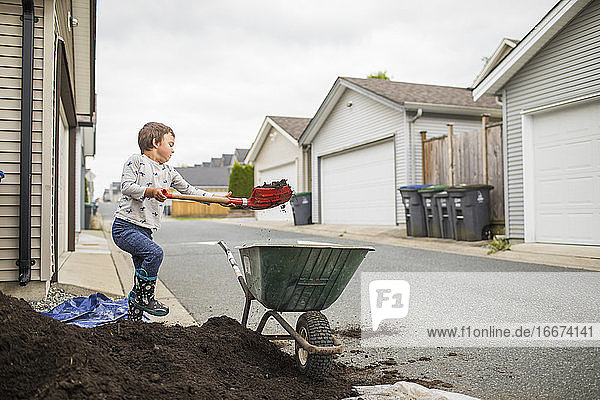 Young boy lifting shovel of dirt into wheelbarrow in back alley