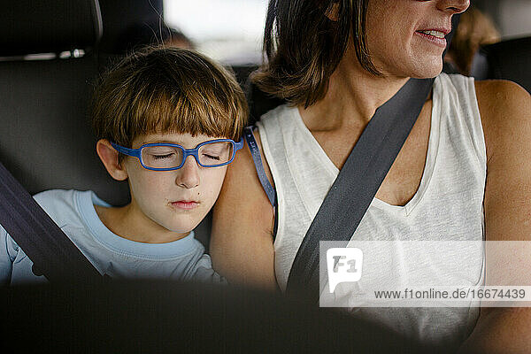 A small child with blue glasses sleeps on mother's shoulder in the car