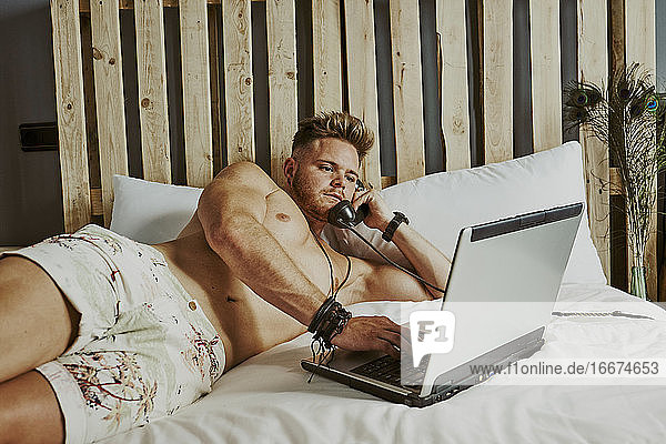 Ein Mann nimmt einen Arbeitsanruf entgegen  während er in einem Hotelbett mit seinem Laptop arbeitet.