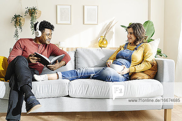 Pregnant wife eating on the couch with her husband who is listening to music and reading a book. Interracial couple concept