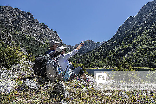Side view of mid aged couple sitting on Spanish Pyrenees mountain and the woman points to the top