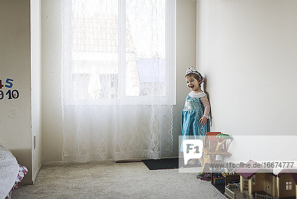 Smiling girl in princess costume standing next to toys by window