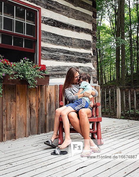 Mutter und Sohn sitzen im Schaukelstuhl auf der Terrasse einer rustikalen Blockhütte.