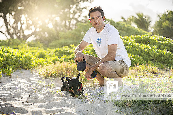 Junger fitter Mann hockt am Strand mit seinem kleinen schwarzen Hund in Hawaii