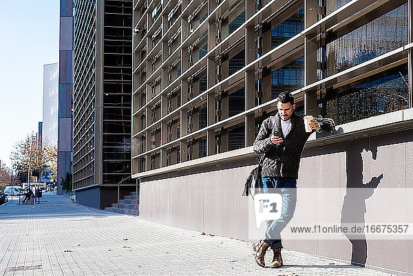 Mann mit Smartphone und Kaffee in der Hand  neben einem Bürogebäude stehend