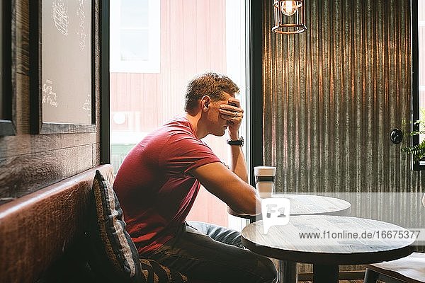 man sat covering his face feeling stressed whilst sat in a cafe