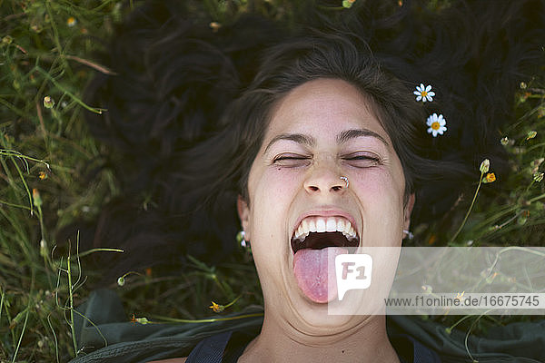 Porträt einer jungen Frau  die im Gras liegt und Blumen in ihrem Haar trägt. Sie streckt ihre Zunge mit einem glücklichen Ausdruck heraus