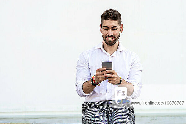 Young bearded man sitting against a white wall using the mobile phone