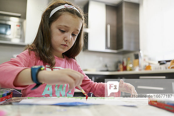 Kleines Mädchen malt eine Zeichnung mit Aquarellfarben in ihrem Haus mit einer Küche im Hintergrund. Kreatives Konzept
