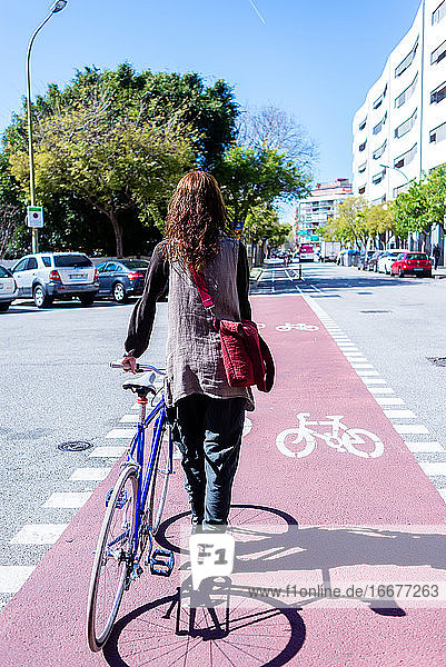 Rückenansicht einer lächelnden Frau  die mit ihrem Fahrrad auf einer Straße in der Stadt spazieren geht