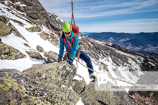 Man climbing rocks above steep snow gully with climbing rope and skis