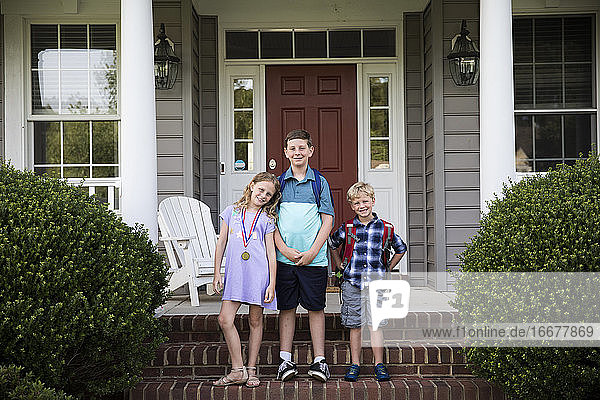 Drei lächelnde glückliche Geschwister mit Rucksäcken stehen auf Ziegelstein Front Steps