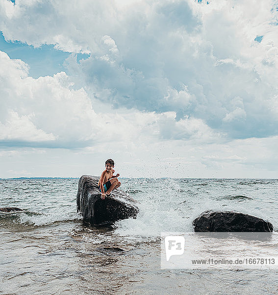 Young boy sitting on big rock in lake getting splashed on summer day.
