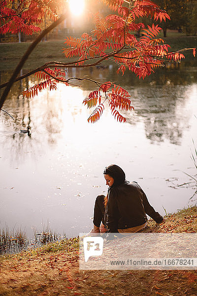 Young woman sitting by lake under a tree during autumn