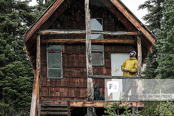 Niedriger Winkel eines männlichen Wanderers  der sich an eine verlassene Holzhütte im Wald lehnt  während er sich beim Trekking in British Columbia entspannt und wegschaut