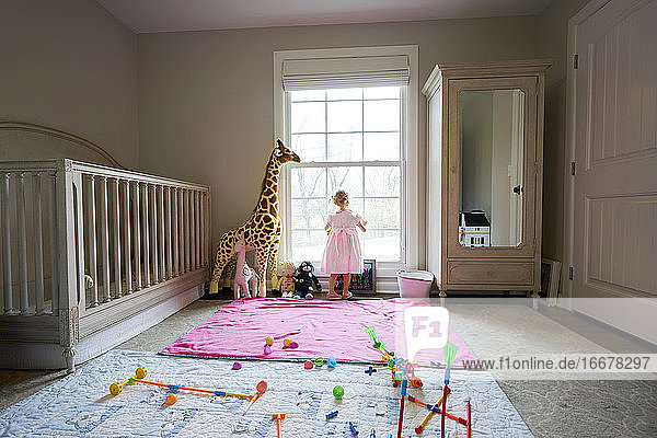 Kleines Mädchen schaut aus dem Fenster ihres Kinderzimmers  Spielzeug auf dem Boden