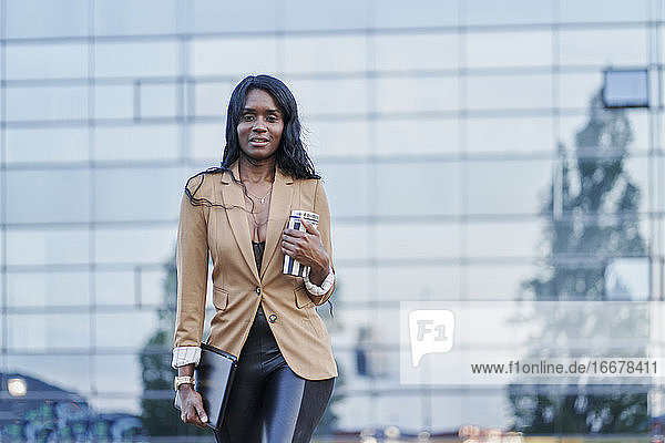 Vorderansicht Porträt einer schwarzen Frau in einem braunen Anzug  die telefoniert und einen Behälter mit Kaffee in der Hand hält