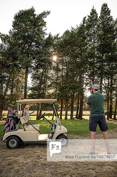 Junges Mädchen mit blondem Pferdeschwanz und Mann mittleren Alters stehen auf einem Golfplatz mit Golfwagen an einem sonnigen Tag.