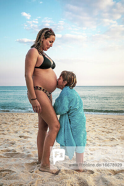 5 Jahre alter Junge küsst den Bauch seiner schwangeren Mutter am Strand