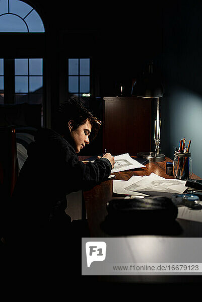Jugendlicher  der an einem Schreibtisch in einem dunklen Raum bei Lampenlicht zeichnet.
