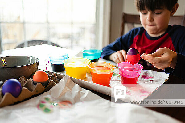 Ein Junge taucht ein Ei in eine Schüssel mit Farbstoff  um es für Ostern zu färben.