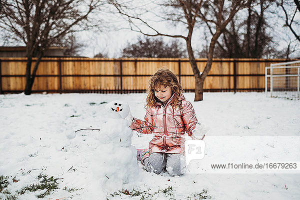 Junges Mädchen sitzt im Schnee und betrachtet glücklich einen Schneemann im Winter