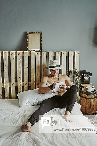 ein junger Mann mit einem Hut  der auf dem Bett in ein Notizbuch schreibt