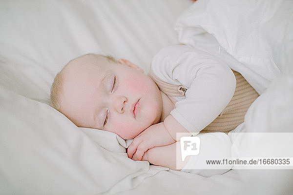 Baby-Mädchen schläft friedlich in weißen Bett mit Blick auf die Kamera mit den Händen für
