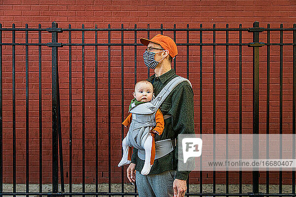 Vater mit Gesichtsmaske trägt seine süße Tochter in der Babytrage und steht während der Coronavirus-Krise am Zaun