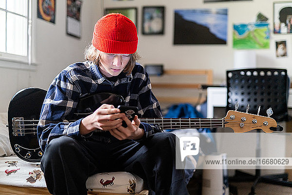 Teenager schaut auf sein Smartphone und hält eine Bassgitarre im Schlafzimmer