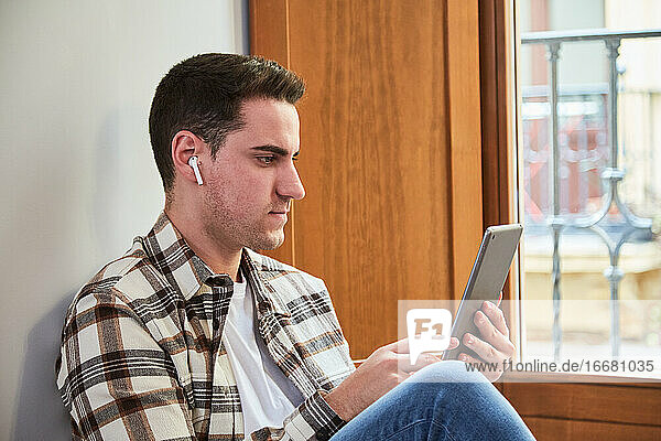 Junger Mann  der zu Hause sitzt und ein digitales Tablet benutzt. Chillen neben dem Fenster beim Konsumieren digitaler Inhalte.