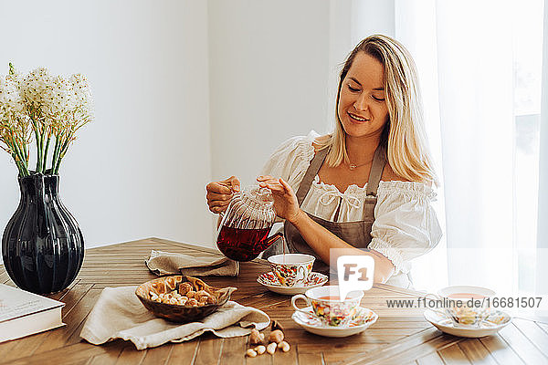 Blonde Frau gießt Hibiskustee in Tassen ein  während sie am Tisch sitzt