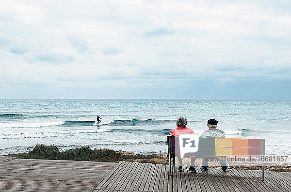 An Old Couple Sitting On A Colorful Bench Looking Out To Sea